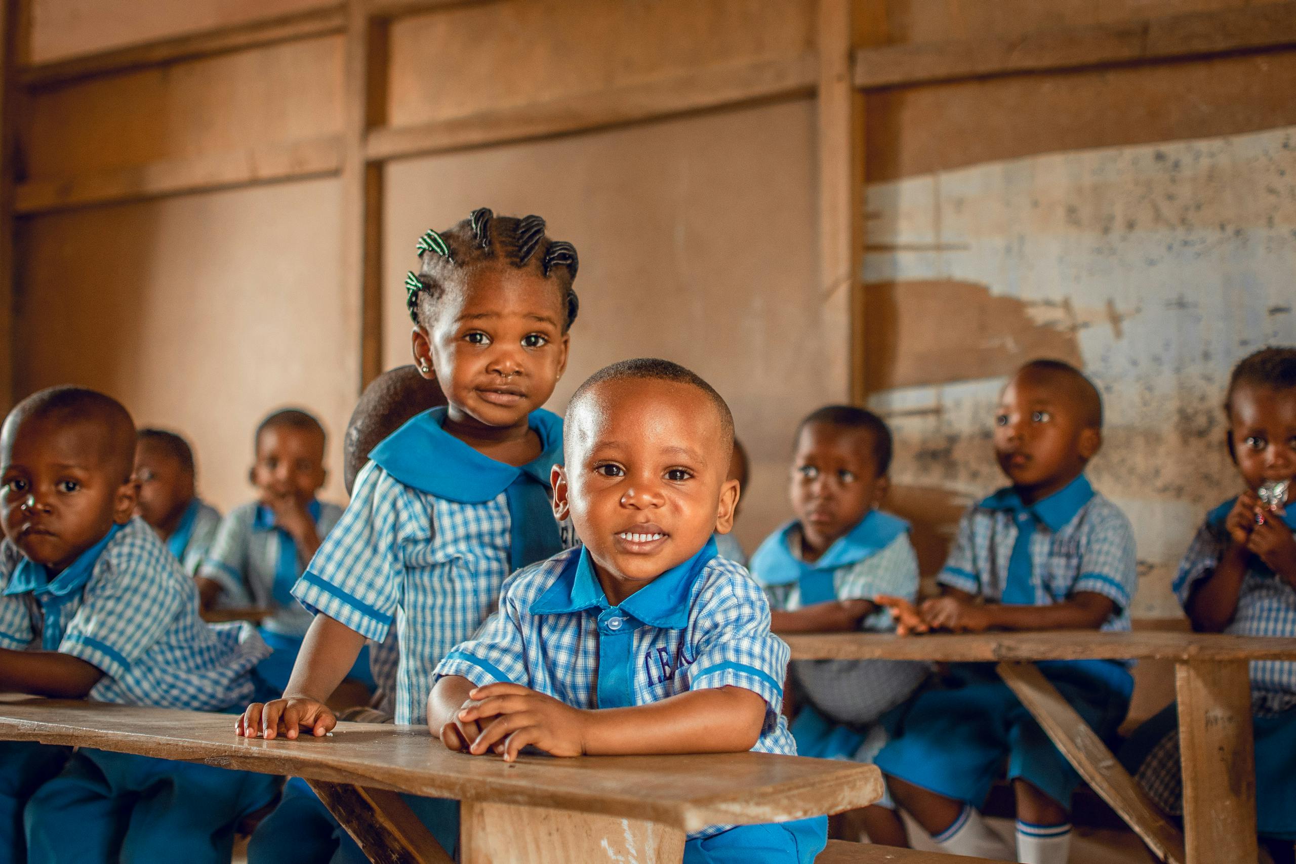 Happy children in a Nigerian classroom wearing uniforms, sitting on wooden benches.