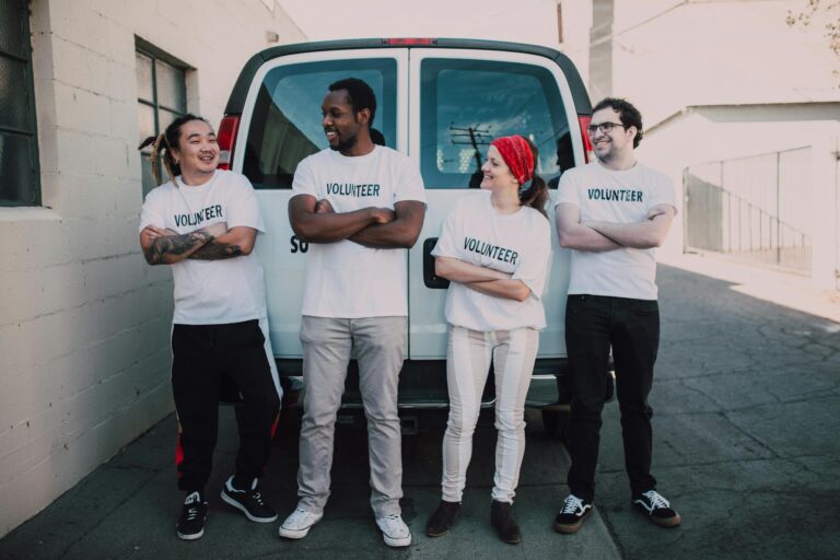 Four diverse volunteers in matching shirts smiling and standing by a van during a community service event.