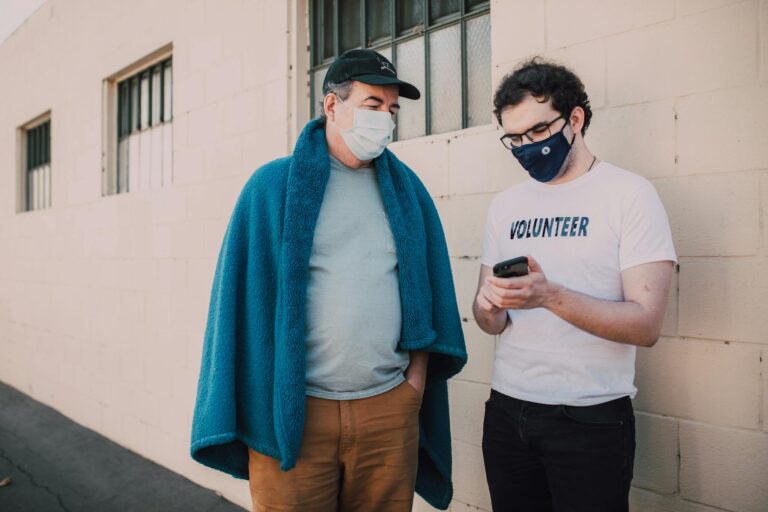 Two masked volunteers stand outside, collaborating on a smartphone project, enhancing community support.